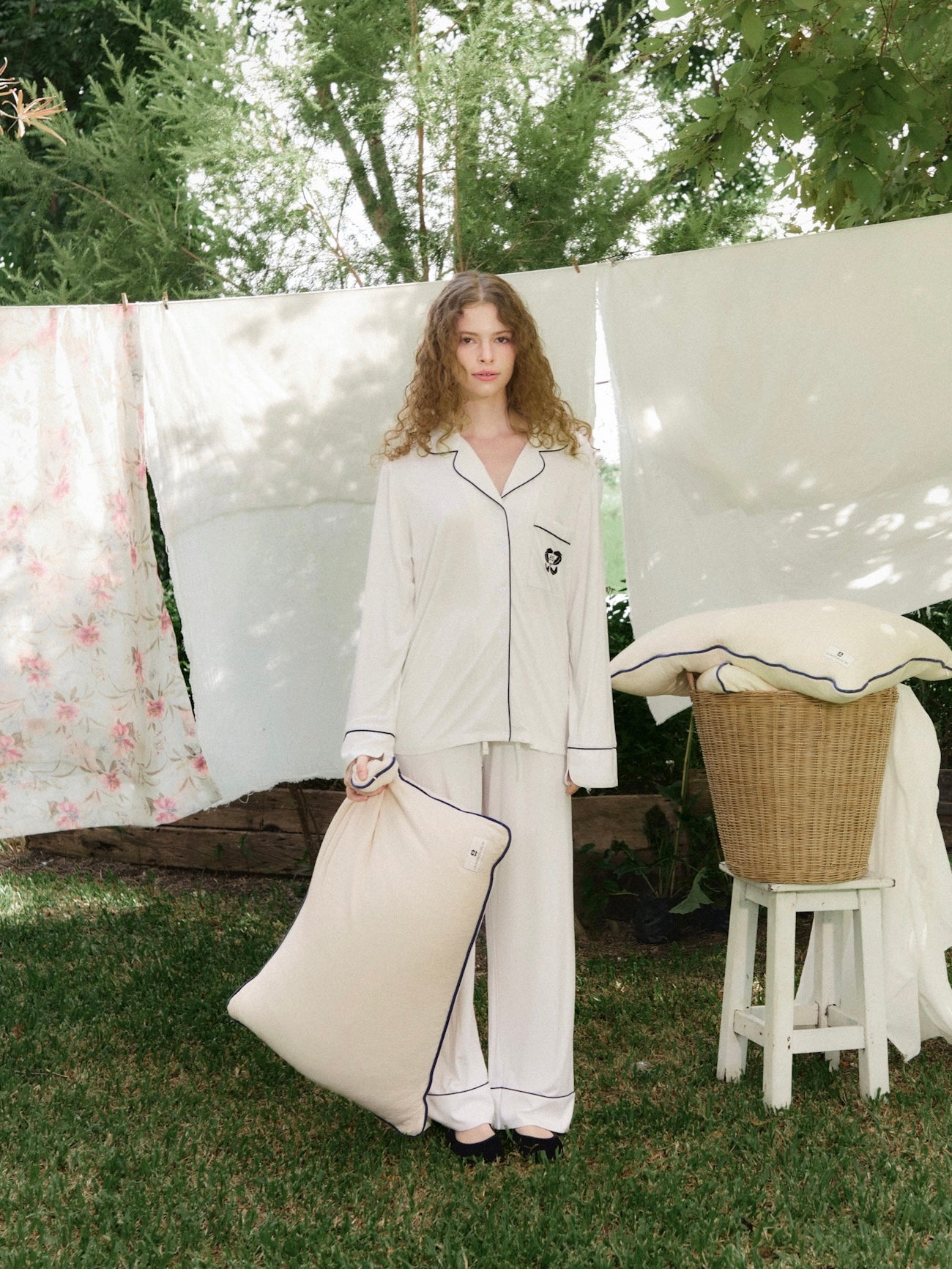 หญิงสาวถือหมอนลาเท็กซ์ออร์แกนิกจาก La Maison du BS ขณะยืนอยู่ในสวนข้างราวตากผ้าผ้าฝ้าย สื่อถึงความสะอาดและความใส่ใจในวัสดุที่ใช้ หมอนผลิตจากวัสดุธรรมชาติ ย่อยสลายได้ biodegradable สาย eco และ vegan-friendly